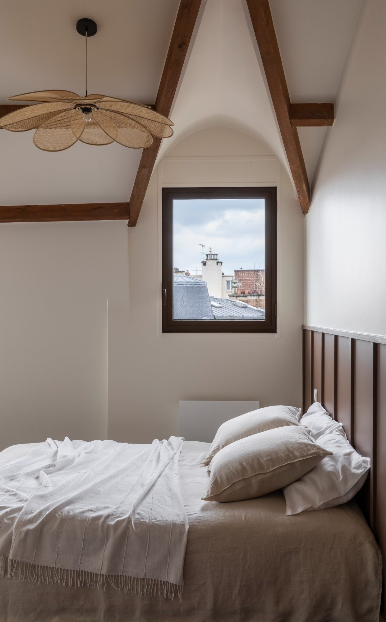 Chambre claire avec lit, tête de lit, fenêtre et poutres apparentes en bois.