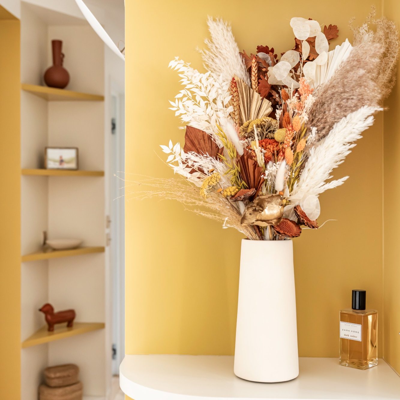Vase de fleurs séchées dans une niche jaune moutarde et décorations simples.