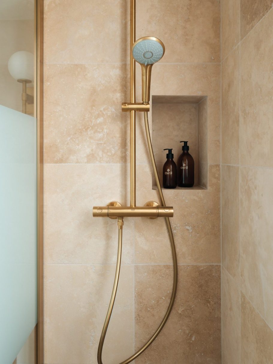 Douche en travertin, avec colonne de douche en laiton et flacons de toilette dans une niche.