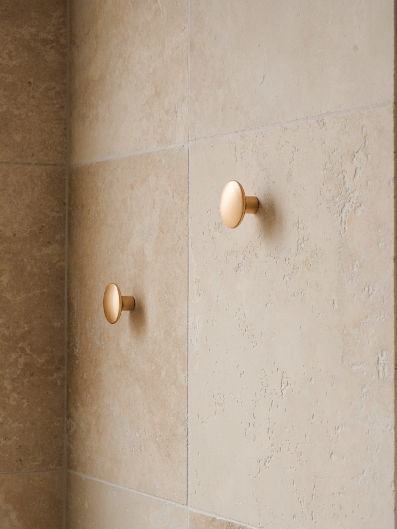 Deux patères dorées sur un mur de travertin beige.