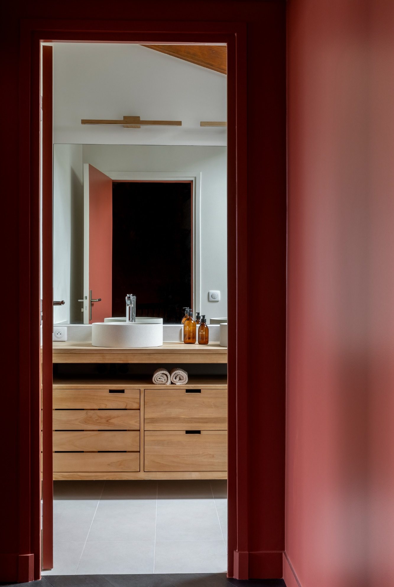 Vue d'une salle de bain moderne avec miroir et meuble en bois depuis un dégagement avec des murs rouge 