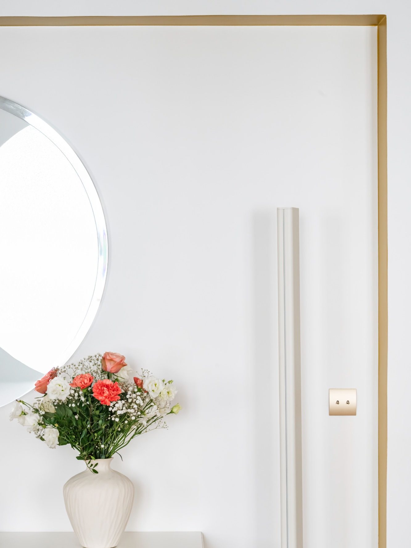 Vase blanc avec fleurs roses et blanches posé sur un bueffet devant un mur blanc avec miroir rond et une arche jaune moutarde