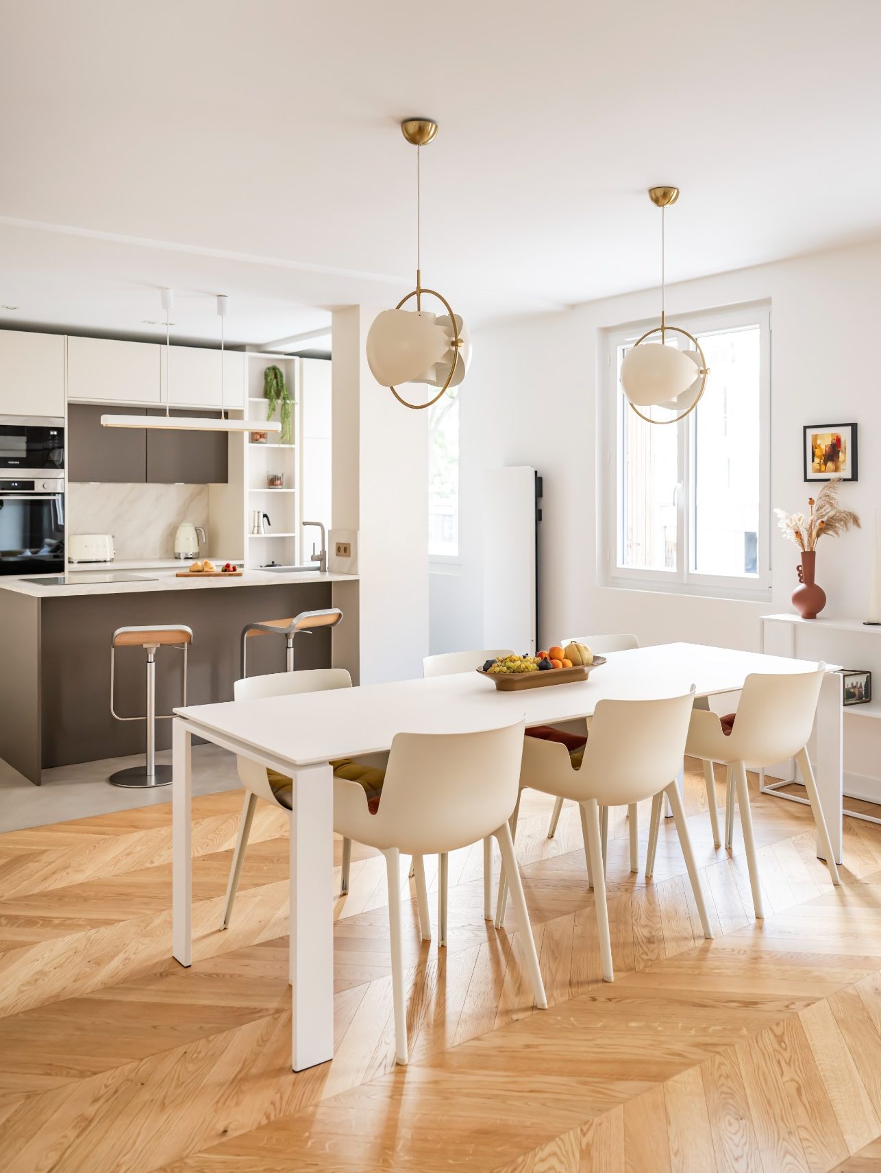 Salle à manger moderne avec une grande table blanche et des chaises, éclairée par des suspensions.