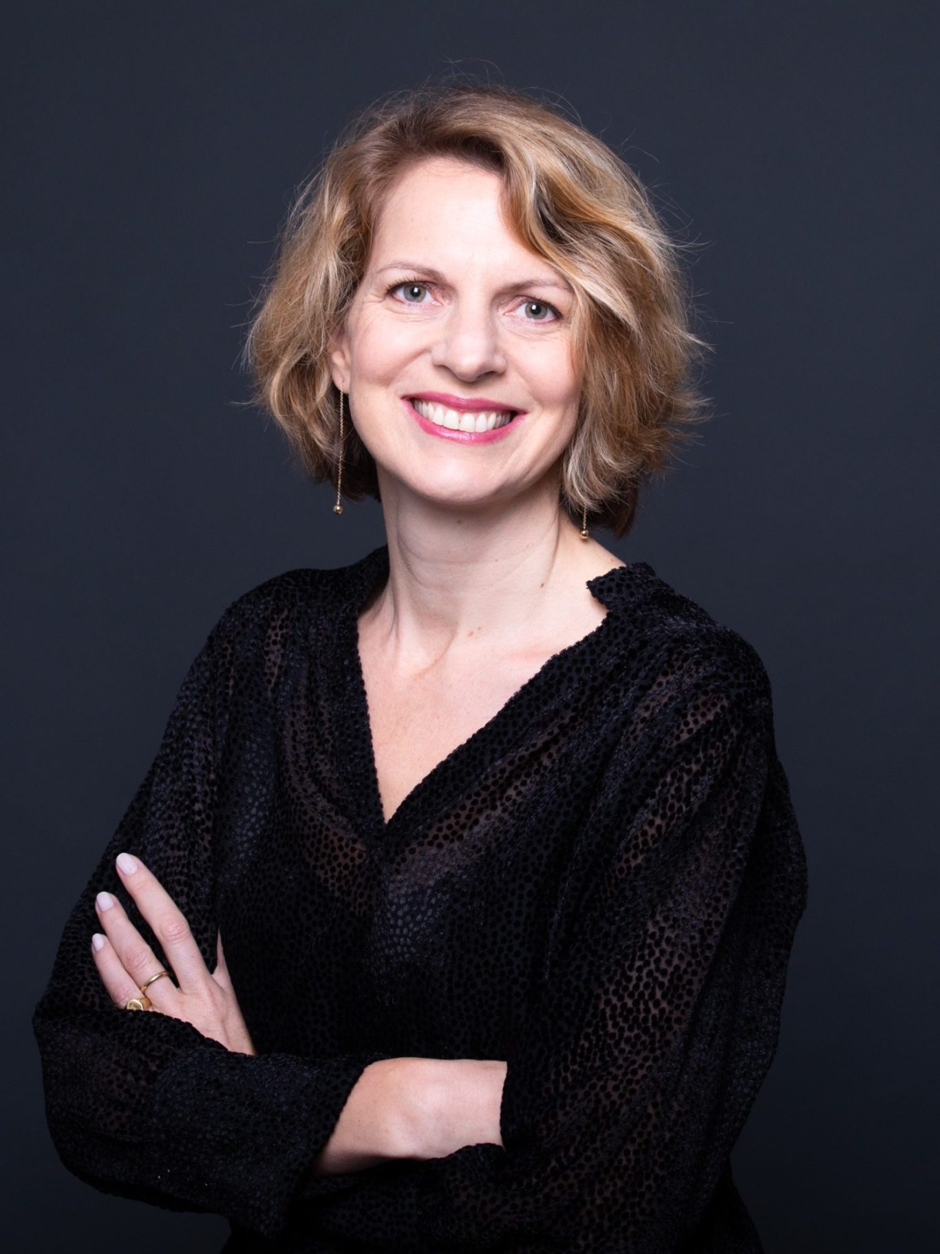 Femme souriante aux cheveux blonds, vêtue de noir, les bras croisés.