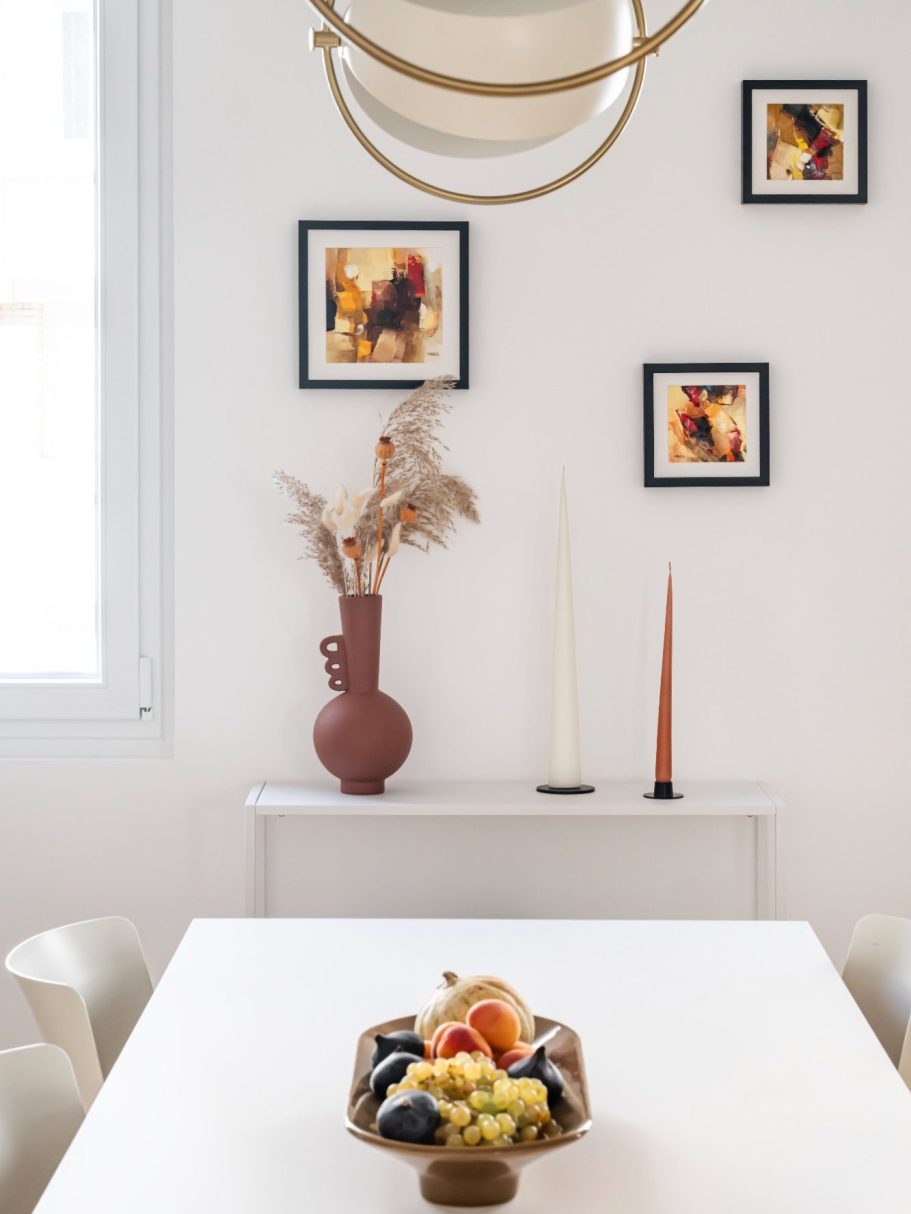 Une salle à manger lumineuse avec un vase, des cadres et un panier de fruits.